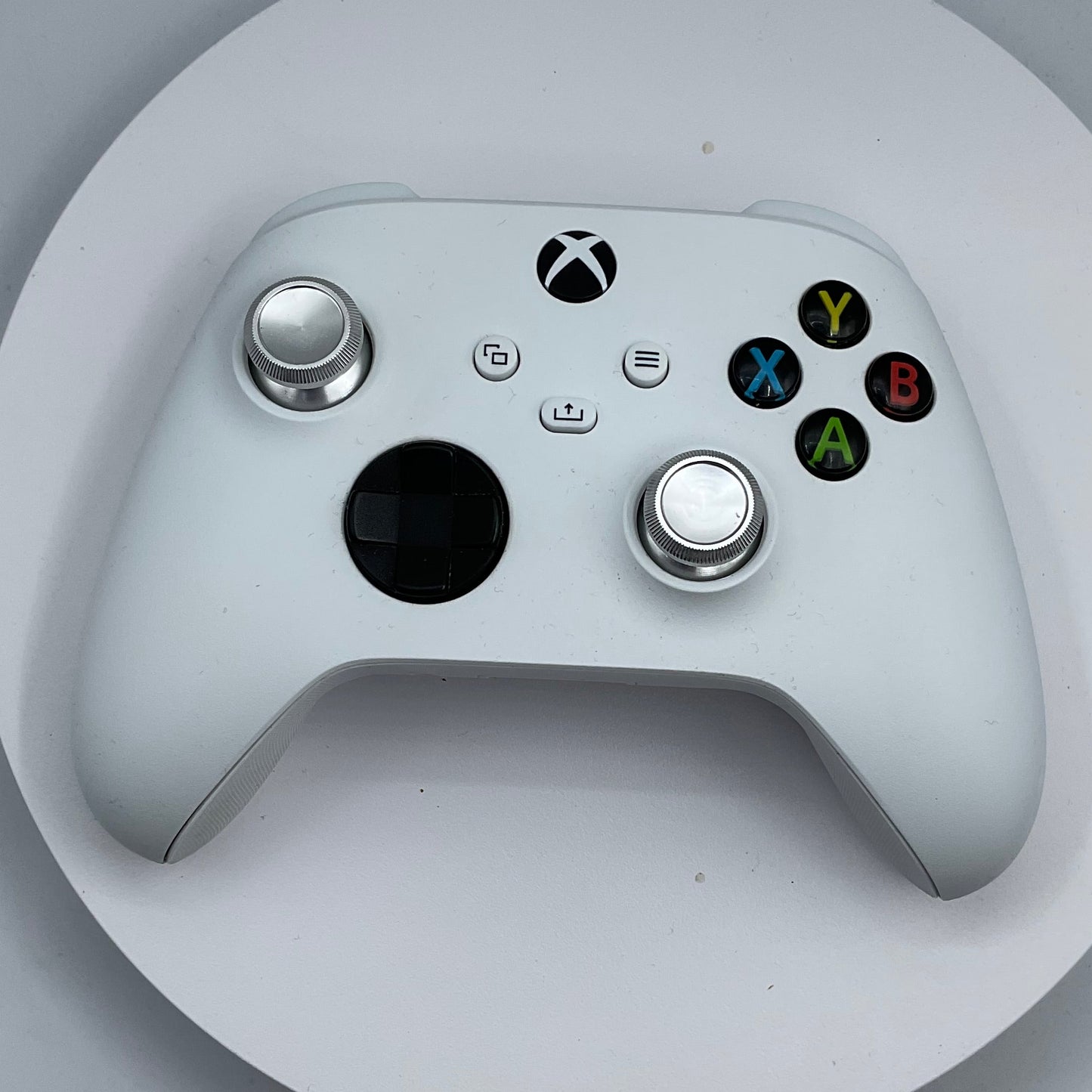 A white Xbox One controller with silver analog sticks and colored face buttons, resting on a round white display stand against a light gray background.