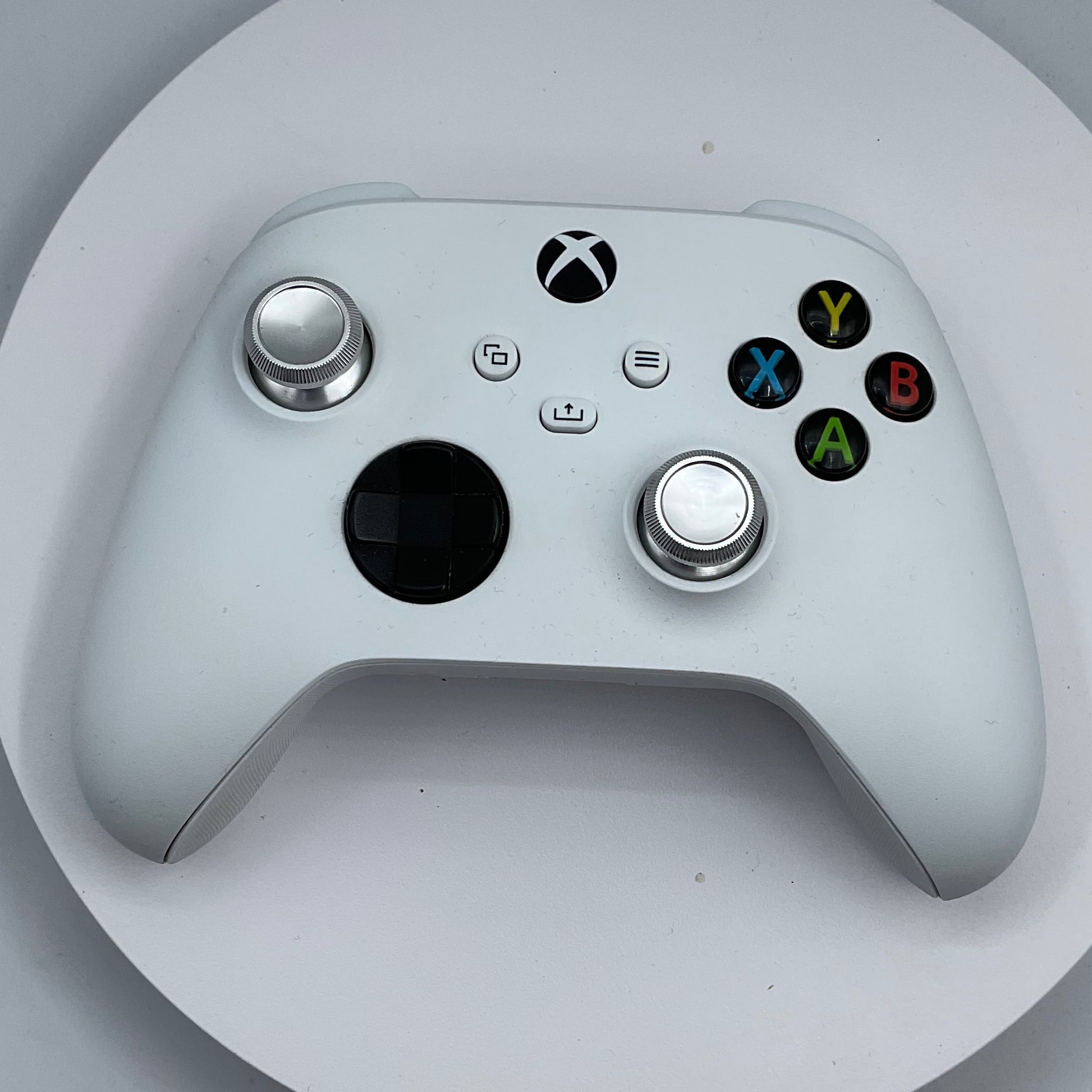 A white Xbox One controller with silver analog sticks and colored face buttons, resting on a round white display stand against a light gray background.